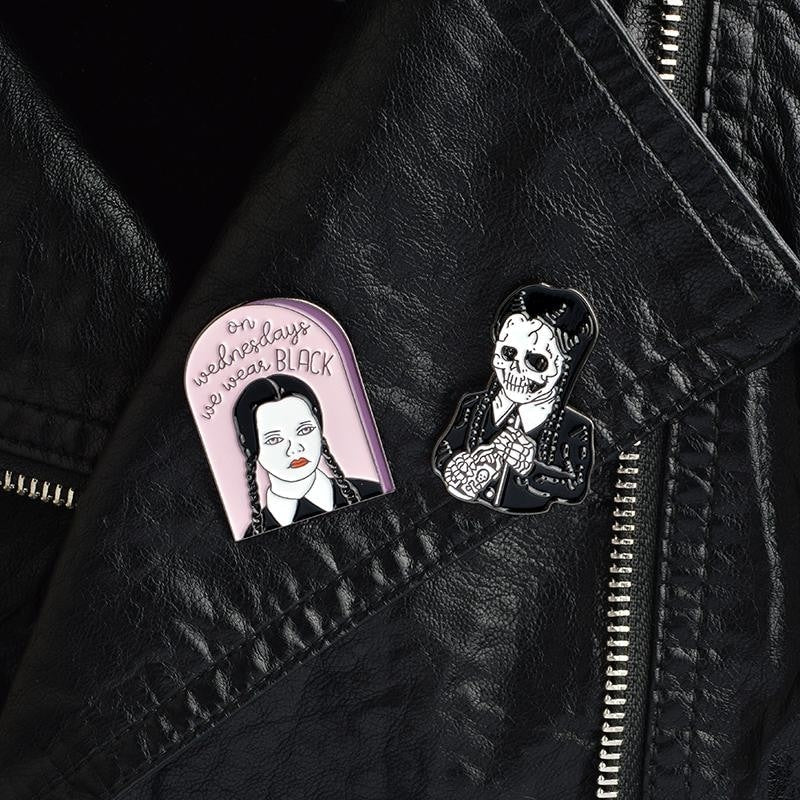 Pink We Wear Black and braided skeleton pins together against a black jacket, with both illustrated fronts facing outward.