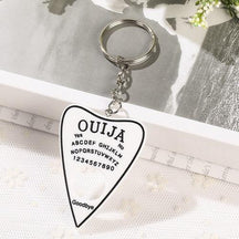 White planchette resin keychain with a black border, alphabet and numbers, resting across an open book on white lace. Its silver-tone ring sits above the short chain.