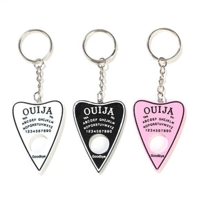 White, black and pink planchette keychains displayed side by side, with contrasting spirit-board lettering and circular openings beneath short silver-tone chains.