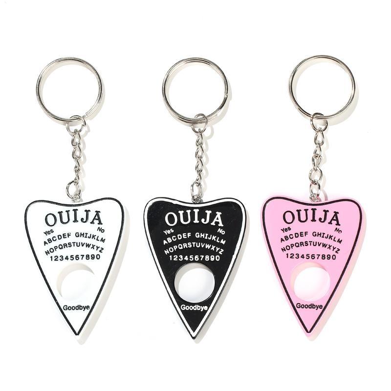 White, black and pink planchette keychains displayed side by side, with contrasting spirit-board lettering and circular openings beneath short silver-tone chains.