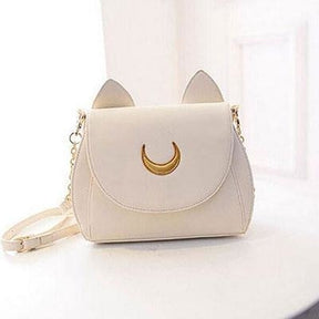 White cat-ear shoulder bag on a pale surface, with a gold-tone crescent at the centre of its curved front flap.