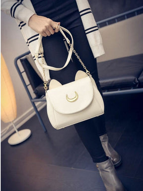 White moon cat bag held by its strap in a standing outfit photo, with the pale front contrasting against dark trousers.