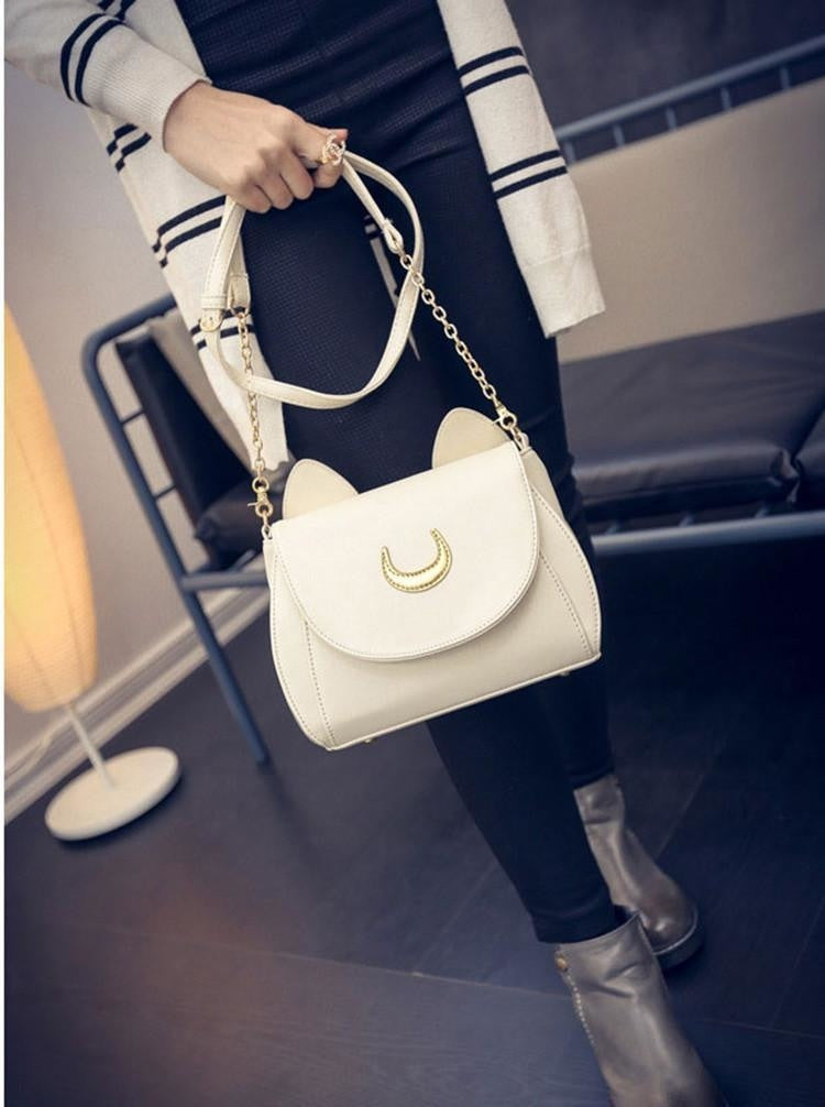 White moon cat bag held by its strap in a standing outfit photo, with the pale front contrasting against dark trousers.
