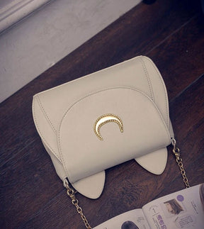 White cat-ear bag lying on a dark wooden surface, showing the curved flap stitching and textured gold-tone crescent.