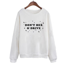 White crewneck sweatshirt on a wooden hanger, with black DON’T HEX & DRIVE lettering surrounded by crescent moons, stars and dots. Long sleeves end in ribbed cuffs above a matching ribbed hem.
