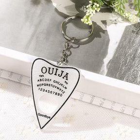 Sparkling white planchette keychain with black lettering and a black border, attached to a short silver-tone chain and ring beside an open book.