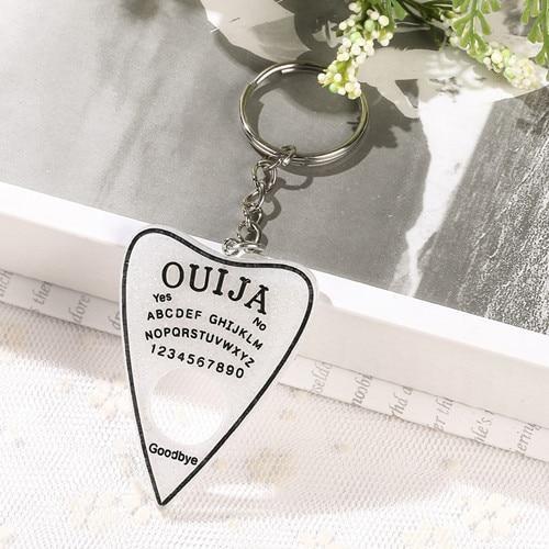 Sparkling white planchette keychain with black lettering and a black border, attached to a short silver-tone chain and ring beside an open book.