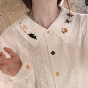 White Halloween blouse worn open at the pointed collar, showing pumpkin, spiderweb and bat embroidery above alternating pale orange and black buttons.