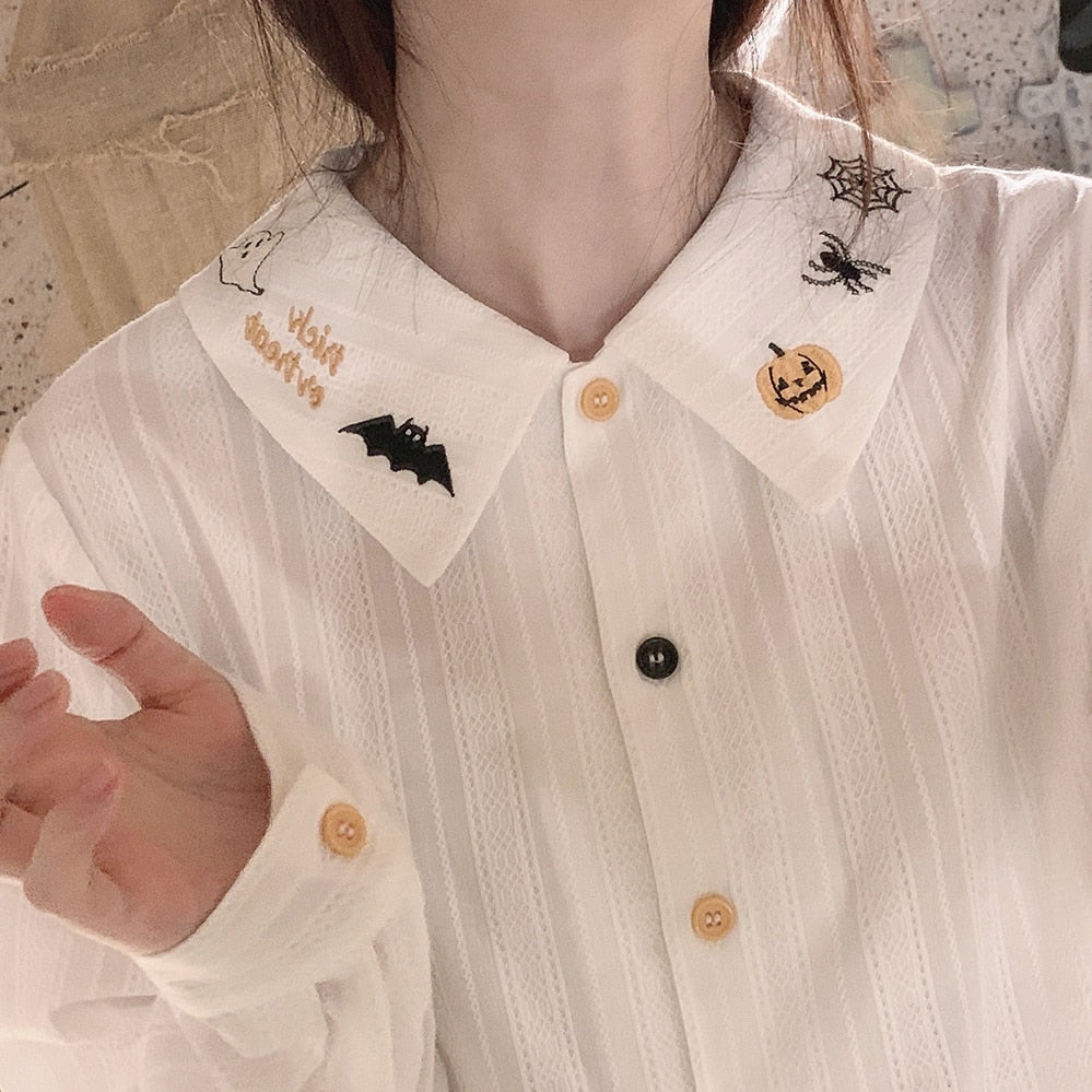 White Halloween blouse worn open at the pointed collar, showing pumpkin, spiderweb and bat embroidery above alternating pale orange and black buttons.