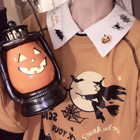 White embroidered Halloween collar layered over an orange witch-print sweatshirt, with a pumpkin lantern held beside the neckline.