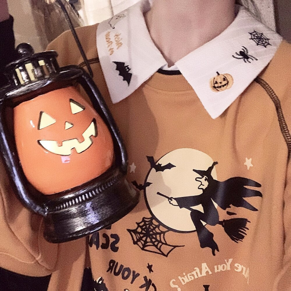 White embroidered Halloween collar layered over an orange witch-print sweatshirt, with a pumpkin lantern held beside the neckline.