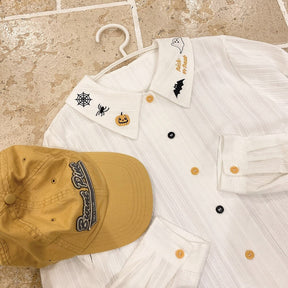 White Halloween blouse laid on a pale brick surface beside a mustard cap, showing its fine vertical texture and embroidered collar.