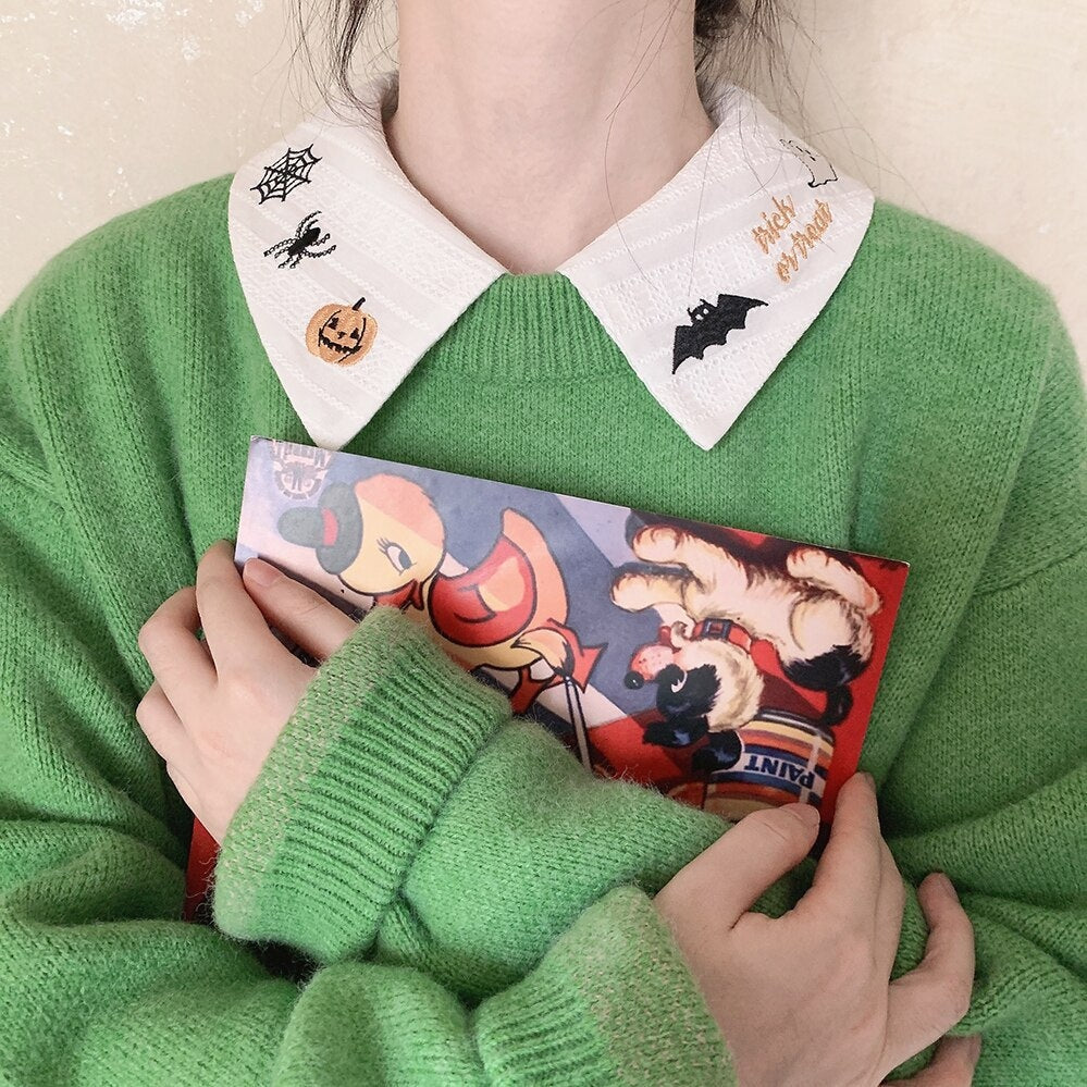 White Halloween blouse collar layered above a green knit cardigan, with a printed card held below the black bat embroidery.
