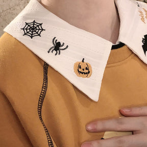 Close detail of the white blouse collar embroidered with a black spiderweb, spider and orange jack-o-lantern above an orange sweatshirt.