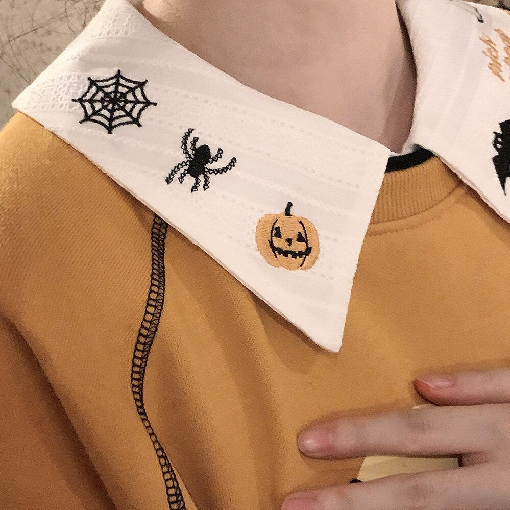 Close detail of the white blouse collar embroidered with a black spiderweb, spider and orange jack-o-lantern above an orange sweatshirt.