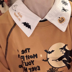Both embroidered collar points of the white Halloween blouse displayed over an orange witch-print sweatshirt, with pumpkin and bat motifs visible.
