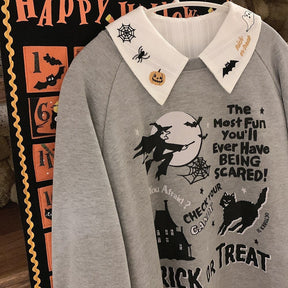 White embroidered Halloween blouse layered under a gray Trick or Treat sweatshirt on a hanger beside an orange Halloween wall graphic.