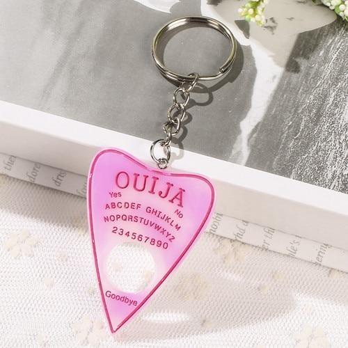 Pale white-to-pink planchette keychain with vivid pink lettering and an outline around the pointed charm, photographed against an open book and white lace.
