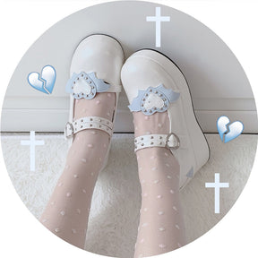 White winged-heart platform Mary Janes worn with sheer white stockings, showing blue heart-and-wing accents beneath decorative cross graphics.