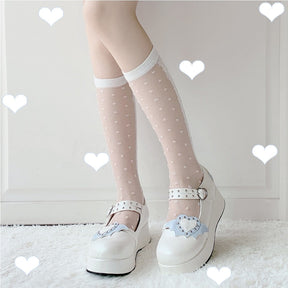 White winged-heart Mary Janes worn with sheer dotted white socks, pictured from the side and front against white heart graphics.
