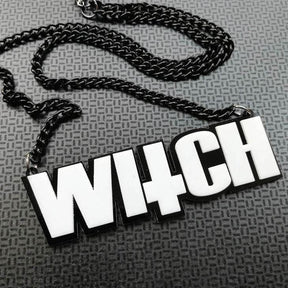 Close view of the WITCH pendant and matching black chain arranged on textured gray fabric. White letter faces contrast with the black outline.