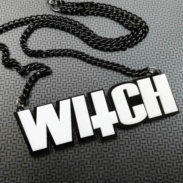 Close view of the WITCH pendant and matching black chain arranged on textured gray fabric. White letter faces contrast with the black outline.