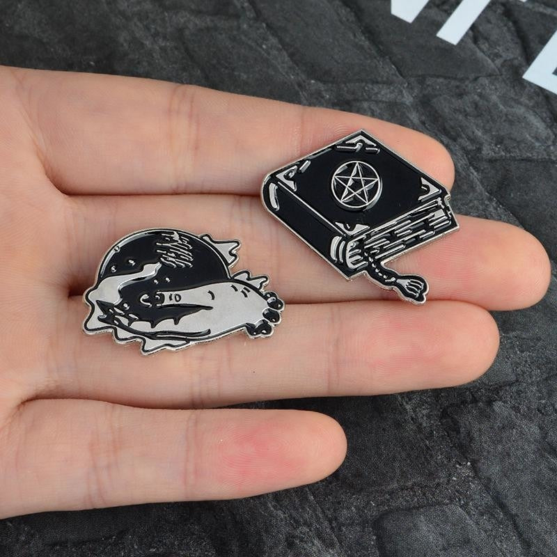 Spellbook and crystal ball enamel pins resting together in an open palm, showing their small scale and black and silver-tone details.