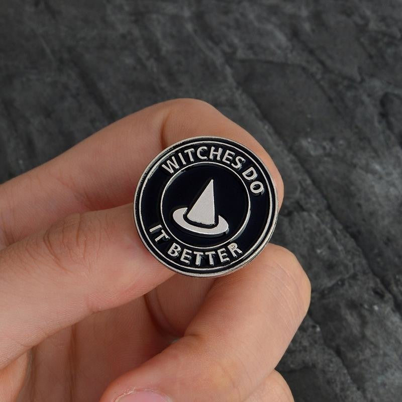 Witches Do It Better pin held between fingers, showing the witch-hat silhouette and circular black-and-silver design.
