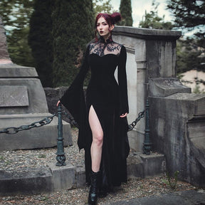 Full-length black velvet gown beside stone monuments, with long pointed sleeves and a slit revealing one leg.