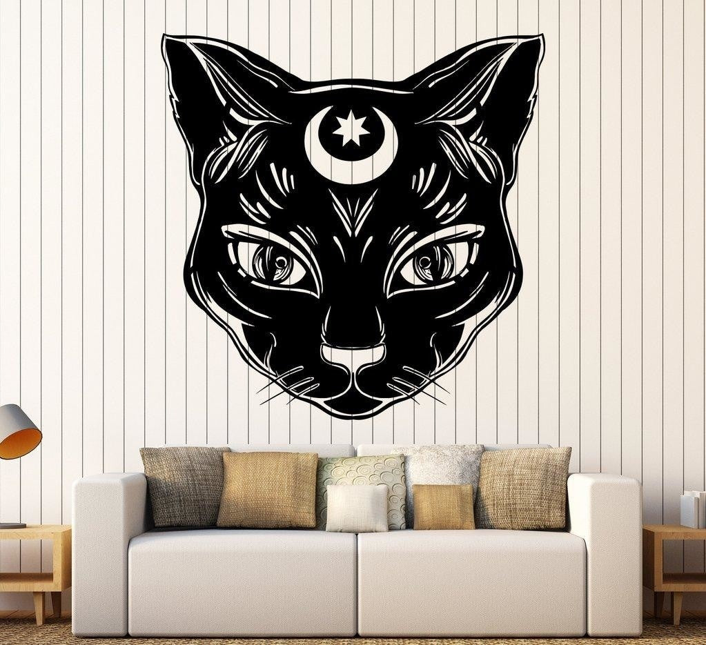 Black witchy cat-face decal displayed above a sofa in a living-room mockup, with detailed eyes and a small celestial forehead motif.