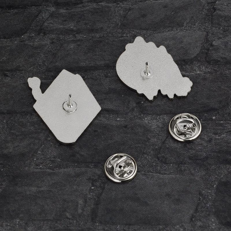 Reverse sides of the spellbook and crystal ball pins, each with a single post. Their removable silver-tone butterfly-clutch backs lie beside them on dark fabric.