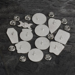 Rear sides of eleven witchy enamel pins arranged on dark fabric, each with a single post and a separate silver-tone butterfly-clutch backing.