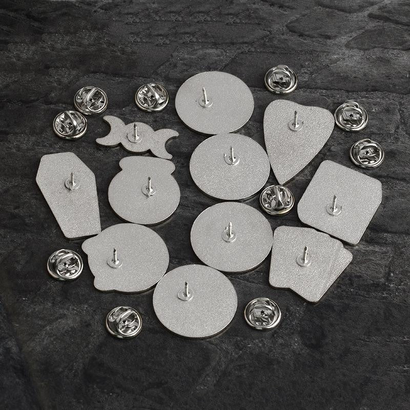 Rear sides of eleven witchy enamel pins arranged on dark fabric, each with a single post and a separate silver-tone butterfly-clutch backing.