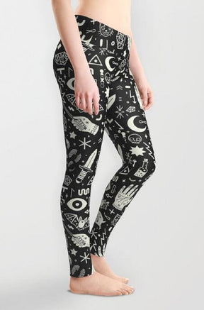 A barefoot model wears black witchy leggings at an angle against a grey background, showing the fitted side profile and white hand, moon and crystal print.