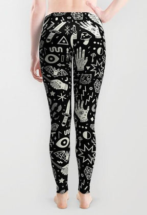 Black occult-print leggings worn by a barefoot model viewed from behind, with white illustrations continuing across the hips and legs against grey.