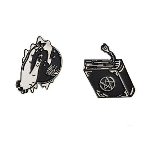 Two black and silver-tone witchy enamel pin designs on white: a hand over a crystal ball and a closed spellbook with a pentacle and tasselled bookmark.