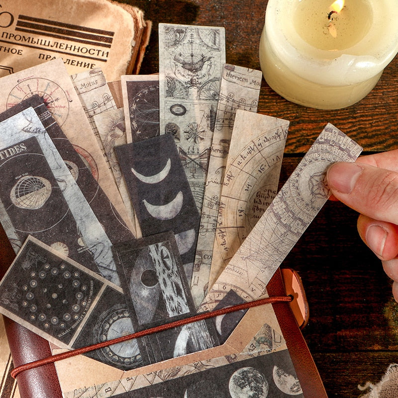 Long moon-phase and celestial stickers fanned from a brown journal beside a candle, with dark lunar illustrations against faded paper colours.