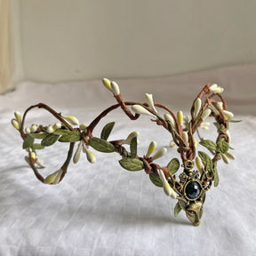 Brown vine-style tiara with pale buds, green leaves and a dark round accent above a deer-head centrepiece.