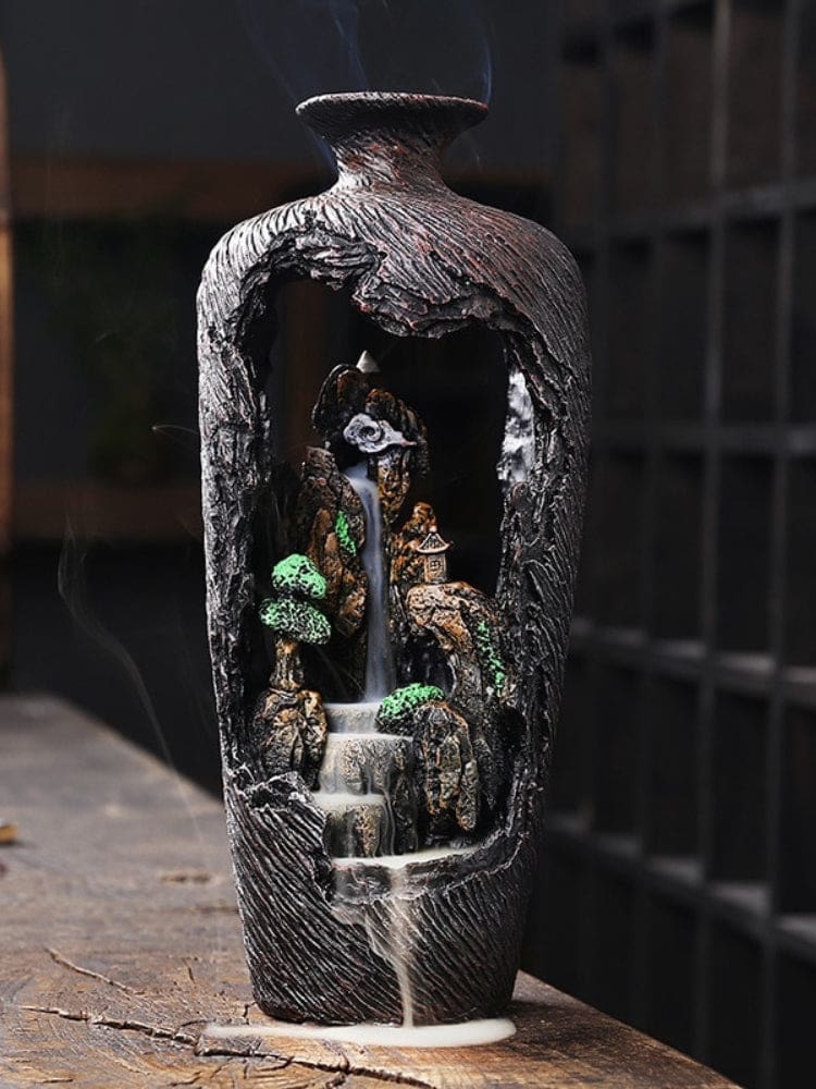 Brown vase-shaped backflow incense burner on a wooden surface, with smoke descending through a miniature rocky landscape and green trees.