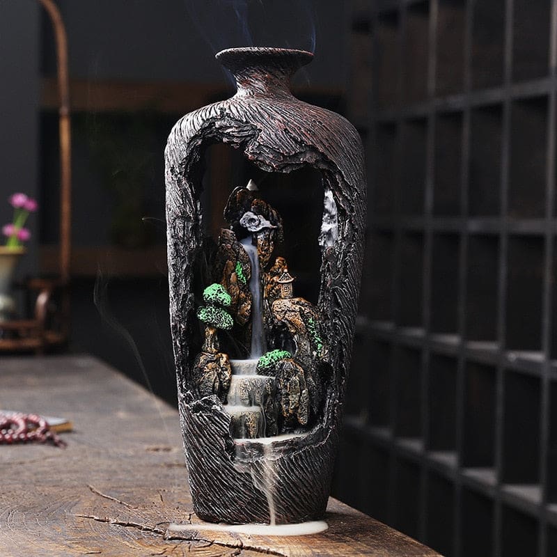 Brown vase incense burner emitting a descending smoke trail on a wooden table beside decorative beads.