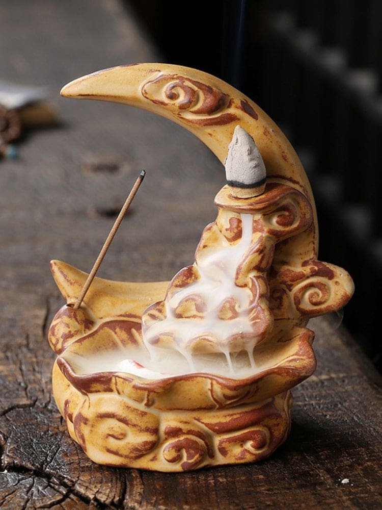 Yellow crescent moon incense burner with warm brown scroll details, shown with a cone and incense stick as smoke cascades into the lower basin.