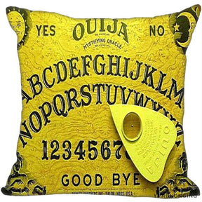 Bright yellow square Ouija cushion cover with dark board lettering, decorative corner illustrations and an angled yellow planchette printed over the alphabet.